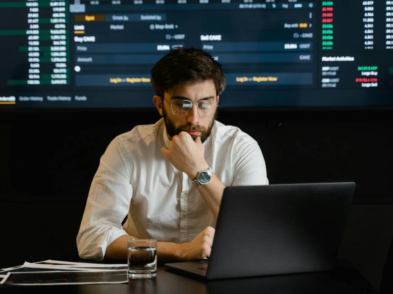 Un señor están mirando el ordenador y por detrás hay gráficos de finanzas en una pantalla